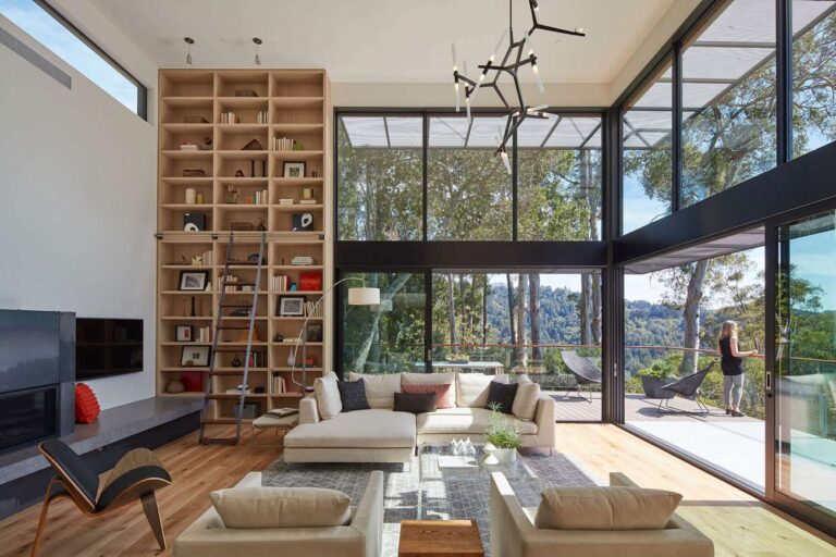 cam duvarlar, tavan yüksekliğinde ahşap kitaplık, krem ​​rengi kanepeler ve koltuklarla geniş oturma odası